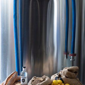 Grand récipient de fermentation en métal avec des tuyaux, des citrons, du pain et une bouteille de vodka Solignac sur une table.