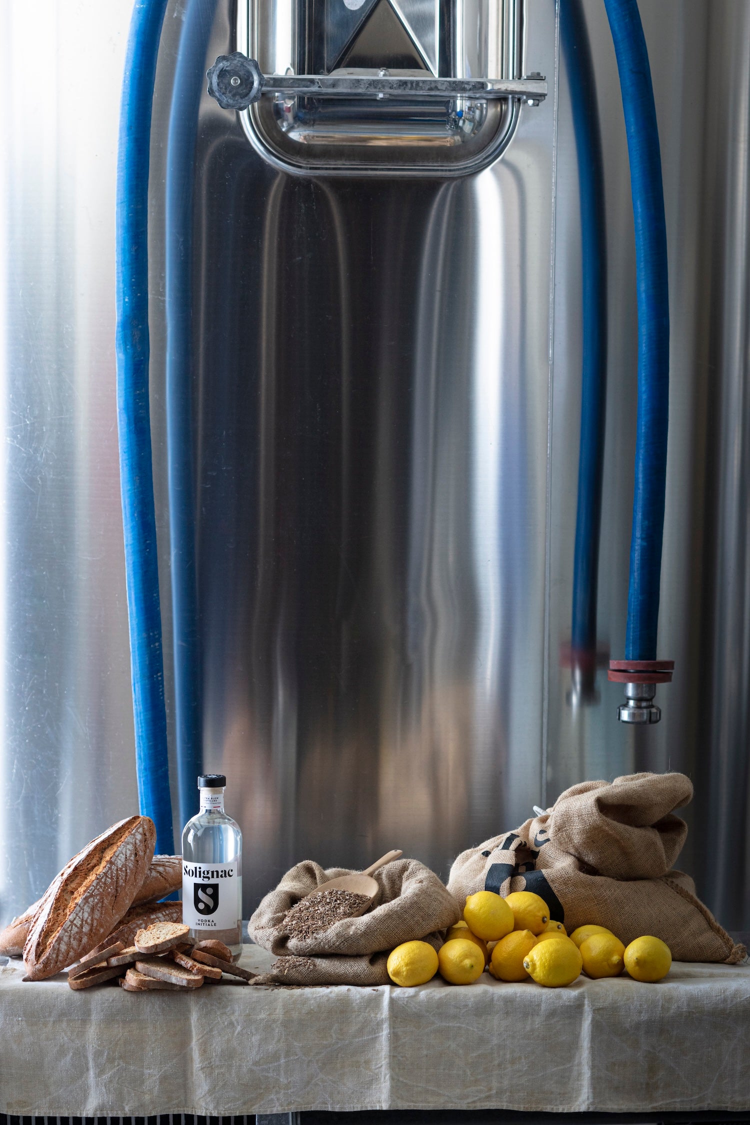 Large metal fermenting vessel with hoses, lemons, bread, and bottle of Solignac vodka on a table.
